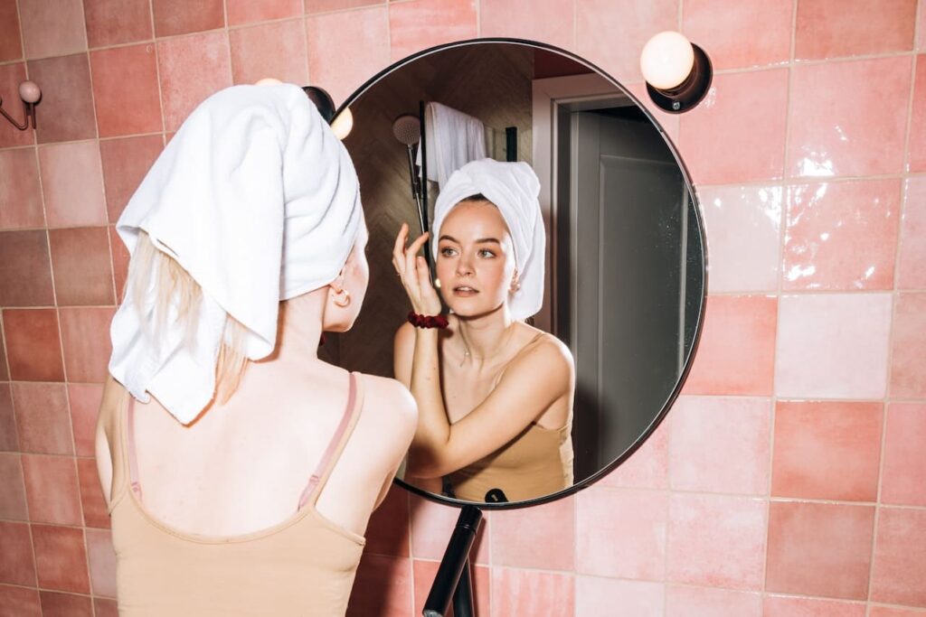 Femme se regardant dans un miroir, serviette sur la t&ecirc;te, salle de bain rose.
